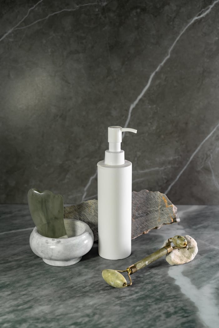 Minimalist skincare arrangement with dispenser, jade roller, and gua sha on marble.